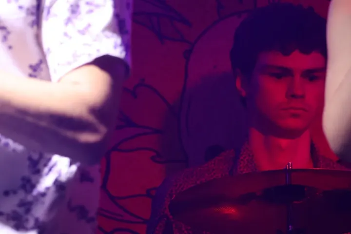 The drummer in purple light, a shoulder blurred in front